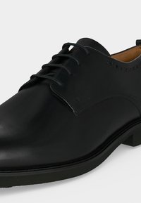 Chaussure formelle Oxford en cuir noir avec laçage fermé, léger détail brogue et talon bas sur fond blanc.