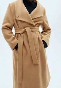 Kabát barvy velbloudí srsti se širokým klopovým límcem, pásem v pase a dlouhými rukávy. Vyroben z měkkého materiálu s texturovaným povrchem.