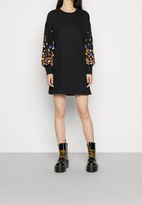 Vestido negro de manga larga con bordado floral colorido en las mangas, combinado con botas de combate negras con cordones. Textura suave en todo el vestido.