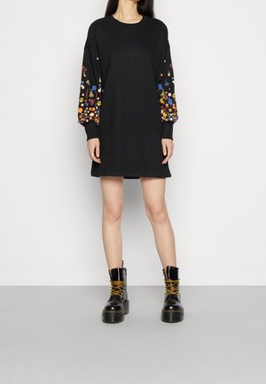 Robe noire à manches longues ornée de broderies florales colorées sur les manches, associée à des bottes de combat noires à lacets. Texture lisse sur l'ensemble.