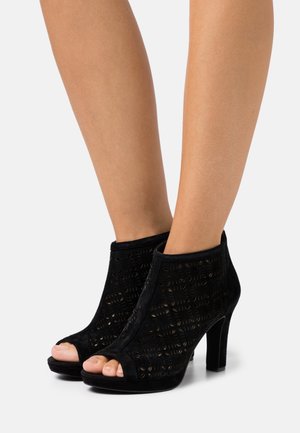 Stopy w czarnych zamszowych botkach na obcasie typu peep-toe z geometrycznymi wzorami i średnim obcasem na białym tle.