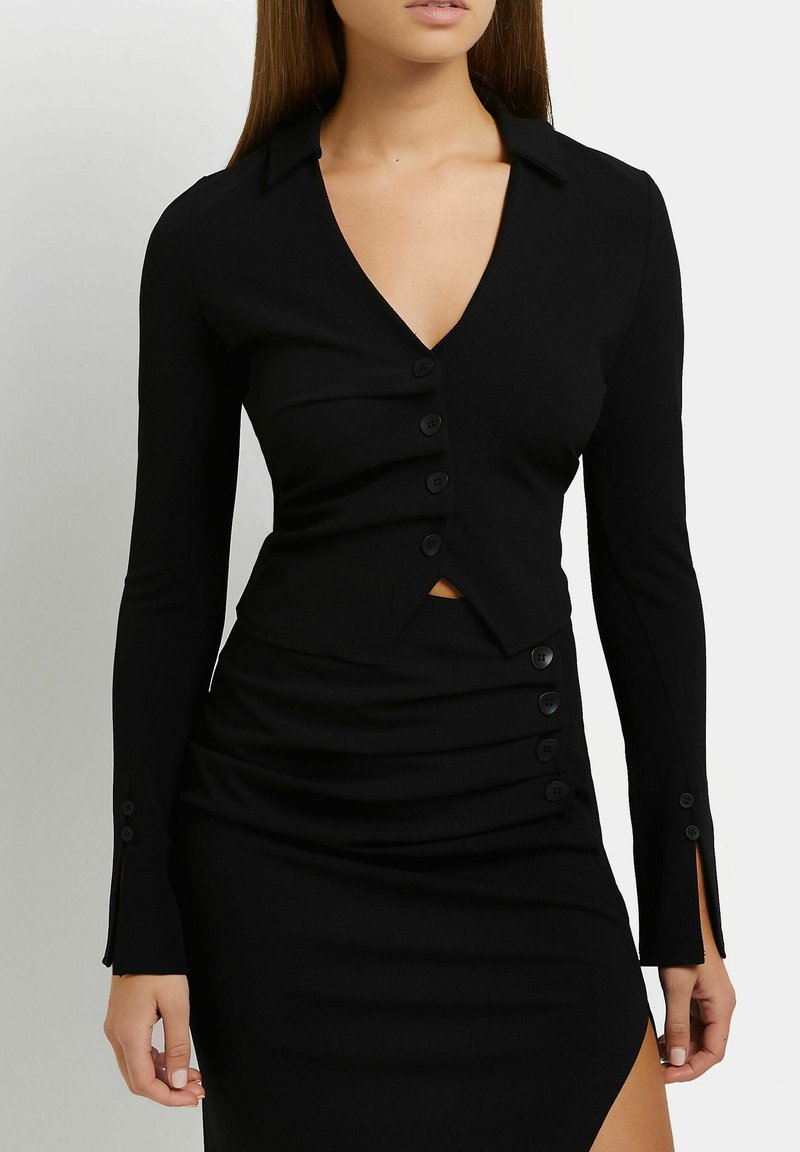 Femme portant un haut noir cintré à manches longues avec col et boutons, et une jupe assortie avec une fente latérale et des détails bouton.