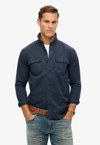 Camisa de manga larga azul marino con cuello alto, dos bolsillos en el pecho, cierre frontal de botones y tejido texturizado. Combinada con jeans celestes y un cinturón marrón.
