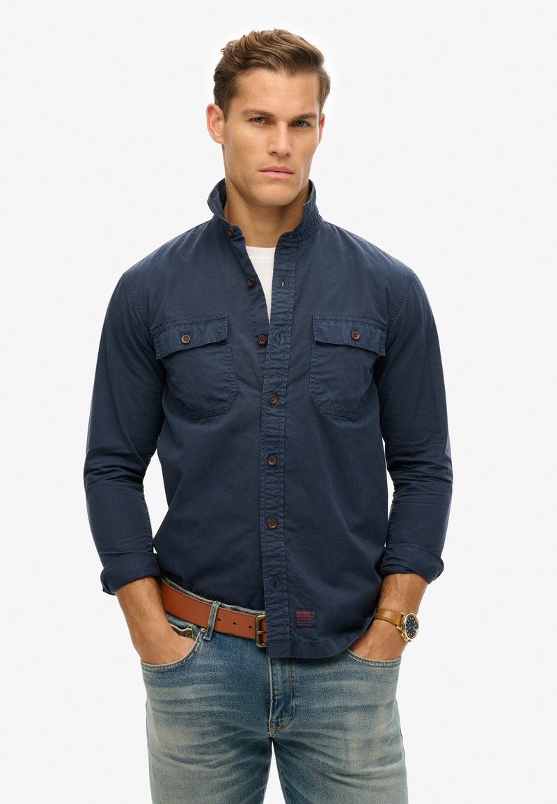 Camisa de manga larga azul marino con cuello alto, dos bolsillos en el pecho, cierre frontal de botones y tejido texturizado. Combinada con jeans celestes y un cinturón marrón.