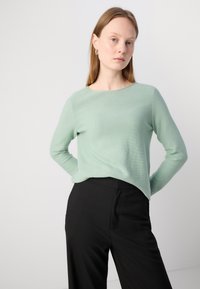 Világoszöld, hosszú ujjú pulóver, kerek nyakkivágással, strukturált anyagból, laza szabásban, fekete, magas derekú nadrággal párosítva.