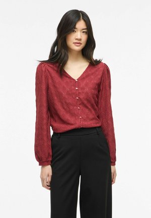 Jeune femme portant un chemisier rouge texturé boutonné avec des manches longues bouffantes et un pantalon noir taille haute sur un fond uni.
