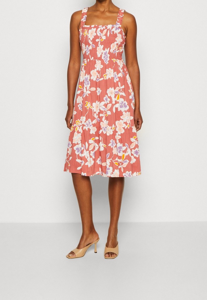 Femme portant une robe corail sans manches, longueur genoux, avec des motifs floraux blancs et violets et des sandales à talons ouvertes beige.
