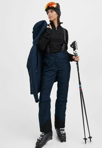Navy ski outfit featuring a zippered top, insulated pants with suspenders, and ski boots. Accessories include a helmet and ski poles.