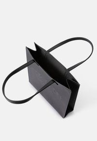 Bolso de mano de imitación cuero negro con dos asas largas, parte superior abierta, nombre de la marca grabado en la parte frontal y diseño estructurado con bordes angulares.