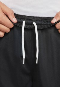 Svarta sportshorts med en texturerad midja, vita dragsnören och en slät tygkvalitet. Närbild av midjeområdet och händer.