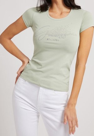 Lichtgroen nauwsluitend T-shirt met korte mouwen. Heeft een zilveren glitter "Guess"-logo met daaronder de tekst "EST. 1981". Gecombineerd met een witte broek.