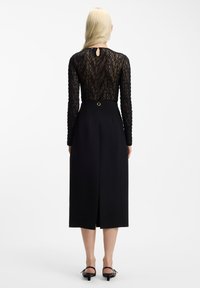 Czarna sukienka z koronkową górą na długie rękawy, detalem w kształcie klucza przy szyi oraz dopasowaną spódnicą z rozcięciem z tyłu i złotym pierścieniem jako ozdobą.