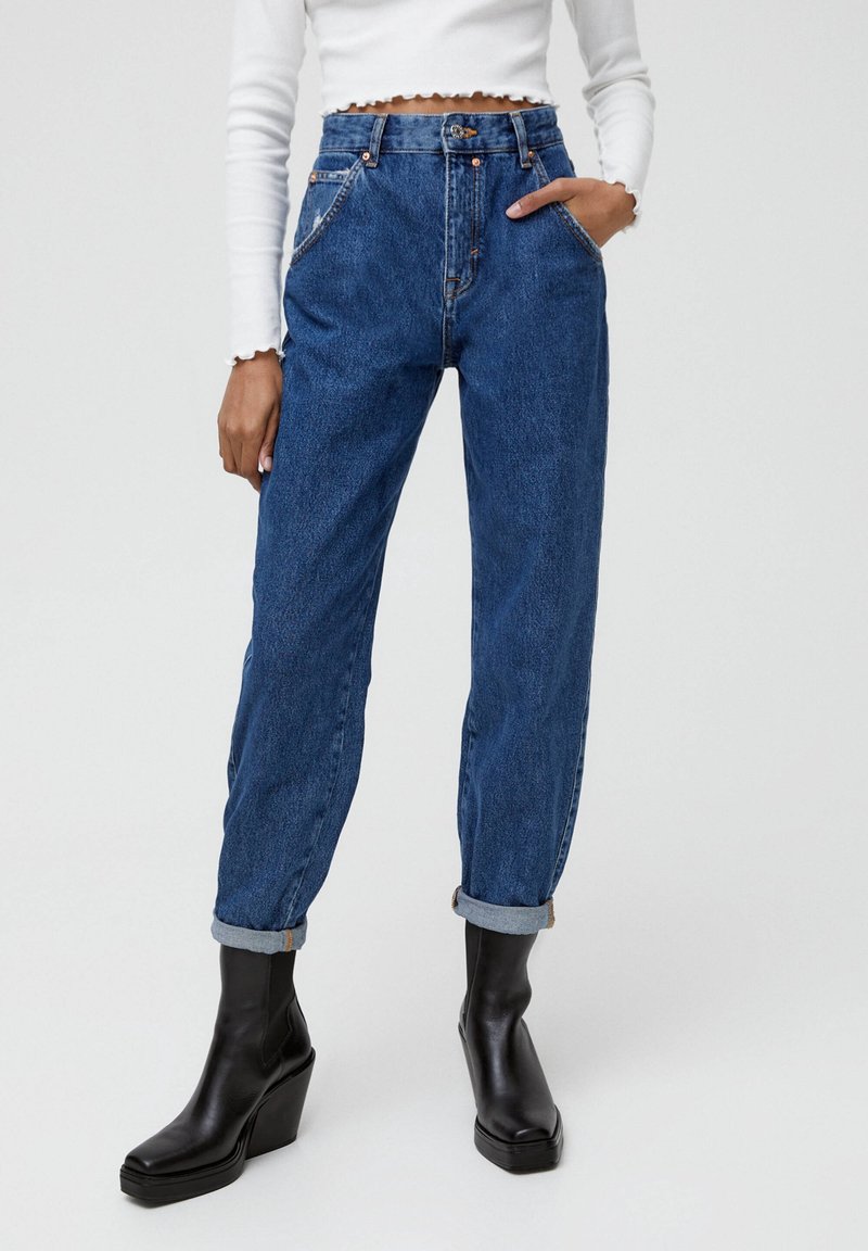 Jeans de mezclilla azul de cintura alta con puños enrollados, bolsillos frontales y costuras en contraste. Combinados con botas de tobillo negras de tacón cuadrado.