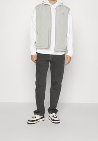 Hellgraue gesteppte Weste über einem weißen Kapuzenpullover, kombiniert mit dunkelgrauen Jeans und gemusterten schwarz-weißen Sneakers. Einfaches Design und lässiger Stil.