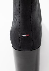 Botín negro de tobillo con un acabado de ante suave, que presenta un tacón alto y brillante en negro y un pequeño logo en rojo, blanco y azul en la parte posterior.