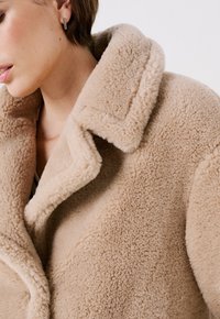 Abrigo de lana beige y esponjoso con un cuello sobredimensionado y costuras visibles. Textura suave con un corte relajado, con un primer plano del tejido.