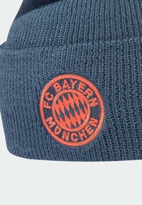 Gorro de punto azul marino con textura acanalada, que presenta un parche circular naranja brillante con "FC Bayern München" y un patrón de rombos en el centro.