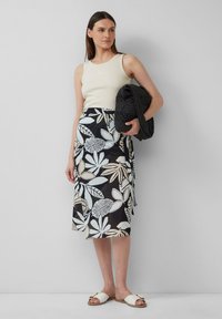 Crèmekleurig geribbeld tanktop, zwarte wikkelrok met grote lichtblauwe en crèmekleurige blaadjespatronen, en witte slidesandalen met gouden accenten.