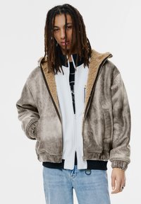 Veste marron en velours avec un col en sherpa beige ; dotée d'une fermeture éclair et de manchons élastiques ; portée par-dessus un sweat à capuche blanc zippé.