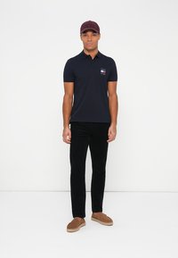 Mørkeblå polo-skjorte med en liten flerfarget logo på brystet, kombinert med svarte bukser og brune loafers, suppleret med en bordeauxrød caps.
