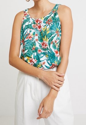 Femme portant un top sans manches à imprimé floral tropical vert, jaune et rouge, associé à un pantalon blanc taille haute.