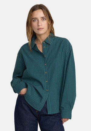 Donna con una camicia ampia a quadri verde e nero con bottoni, una mano nella tasca dei jeans blu scuro su sfondo bianco.