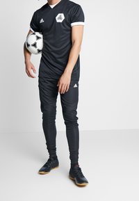 Conjunto atlético de fútbol negro con escote en V, acentos blancos en las mangas y pantalones, con un logotipo hexagonal. El jugador sostiene un balón de fútbol.