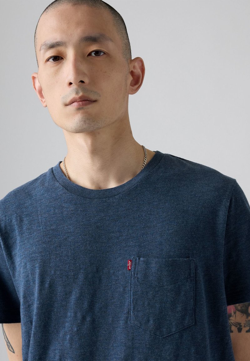 Mann mit rasiertem Kopf, trägt ein blaues Levi’s T-Shirt mit Brusttasche und silberner Kettenhalskette, sichtbares Tattoo am linken Arm vor grauem Hintergrund.