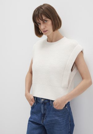 Cremfarbener, gerippter, ärmelloser Pullover mit Rundhalsausschnitt und seitlichen Schlitzen, kombiniert mit blauen, hoch taillierten Denim-Jeans.