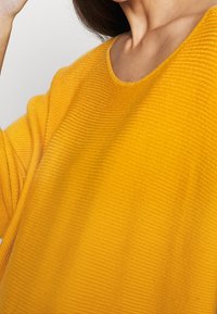 Femme portant un pull en maille jaune moutarde texturé, montrant le décolleté et une partie du bras, sur un fond clair uni.