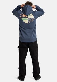 Marineblauwe hoodie met een grafisch ontwerp op de achterkant, met abstracte vormen in perzik, groen en crème. Geribbelde manchetten en zoom. Zwarte cargobroeken.