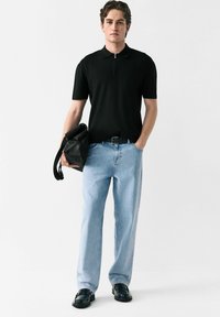 Sort kortærmet polo med lynlås krave, lyseblå lige ben jeans, og sorte lædersko. Mand holder en sort taske.