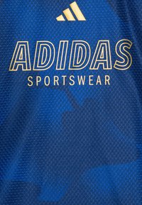Modrý sportovní dres z texturované látky s zlatým logem "ADIDAS" a nápisem "SPORTSWEAR", na pozadí geometrického vzoru.