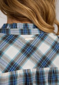 Chemise à carreaux présentant des motifs bleus, gris et blancs, avec un détail de col froncé et une étiquette de marque métallique à l'arrière. Texture de tissu lisse.