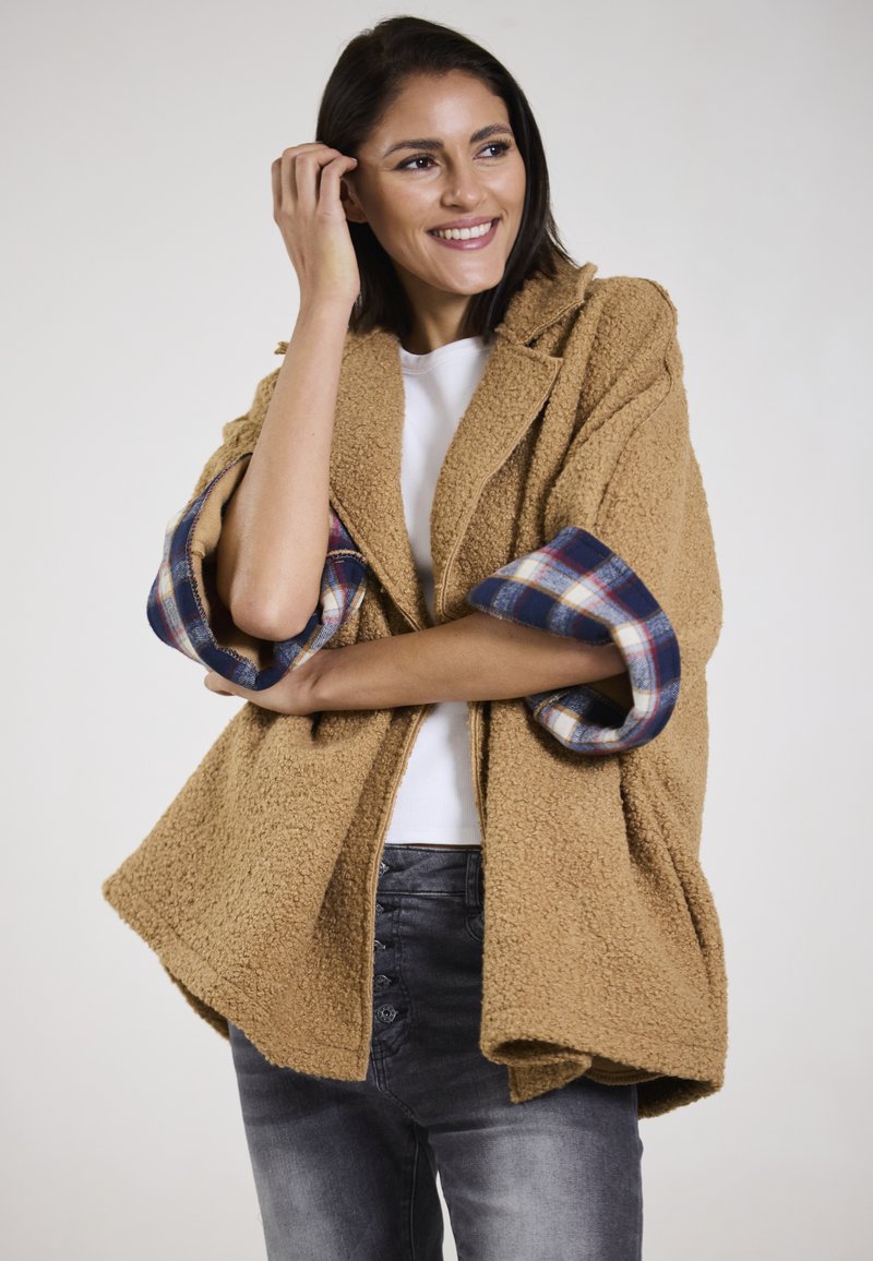 Oversized tan fleece jacket with rolled plaid cuffs, featuring a notched collar and open front design. Paired with dark jeans.