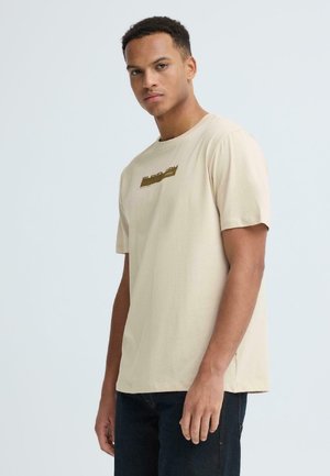 Man draagt een beige T-shirt met korte mouwen en een klein gouden logo, staand tegen een effen lichtgrijze achtergrond.