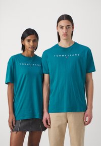 Tehasehe sinise rohelise tooniga lühikeste varrukatega T-särgid, millel on "TOMMY JEANS" logo. Üks mudel kannab teksaseelikut; teine kannab heledat värvi pükse. Lihtne ja mugav disain.
