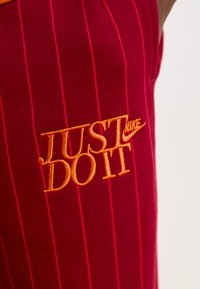 Red striped athletic shorts featuring orange "JUST DO IT" Nike embroidery on the left side. Made from soft fabric with a smooth texture.