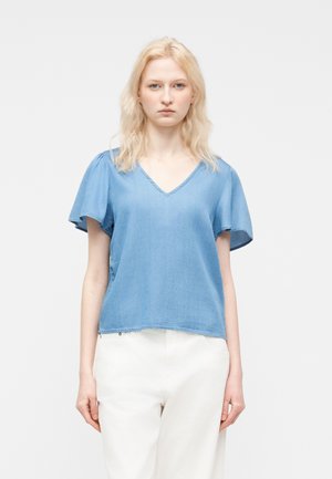 Młoda osoba o jasnych włosach, ubrana w jasnoniebieski krótki rękaw z dekoltem w serek i białe spodnie, stojąca na jednolitym tle.