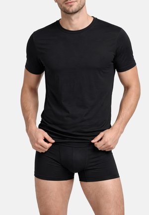 Uomo che indossa una maglietta nera aderente a maniche corte e boxer neri, tenendo il bordo inferiore della maglietta con entrambe le mani.