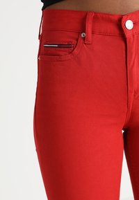 Acercamiento a la parte media con jeans de mezclilla rojos brillantes con un pequeño detalle de tela a rayas en el bolsillo delantero y cierre de botón plateado.