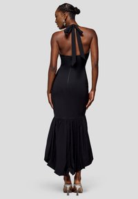 Sort halterneck-kjole med tætsiddende korset og en udvidet, flæset kant. Glat stoftekstur med et bundet sløjfedetalje bagpå.