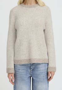 Beżowy sweter w paski z prążkowaną teksturą, okrągłym dekoltem i kontrastującym, jednolitym wykończeniem dolnym. Połączony z jasnoniebieskimi dżinsami.