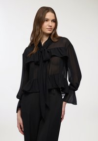 Blusa nera trasparente con scollatura annodata, design a volant, maniche lunghe e tessuto testurizzato, abbinata a pantaloni neri sartoriali.