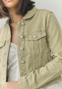 Light olive green denim jacket with a collared neckline, silver snap buttons, and two chest pockets. Worn over a white lace-trimmed top.