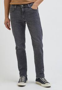 Vaqueros de mezclilla gris con corte recto y un ligero efecto de desgaste. Presenta bolsillos frontales, cremallera y detalles de remaches. Combinados con zapatillas negras.
