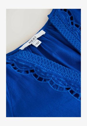 Plava bluzica s raskošnom izrezanom vezom uz izrez. Izrađena od glatkog materijala s opuštenim, lepršavim oblikom.