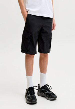 Persoon die zwarte cargoshorts, witte sokken, zwarte sneakers en een wit t-shirt draagt met handen in de zakken, staand op een witte achtergrond.