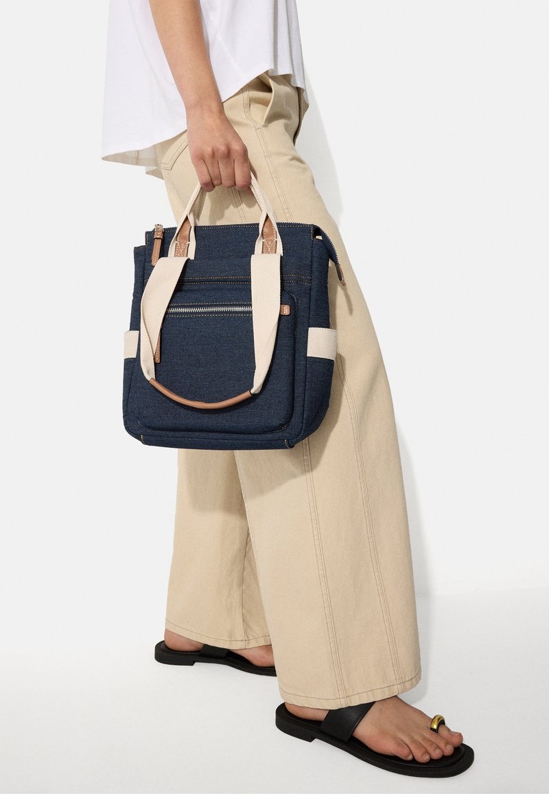 Sac fourre-tout en denim bleu marine avec des accents en tissu crème, deux anses supérieures et une poche zippée à l'avant. Le design présente une forme structurée.