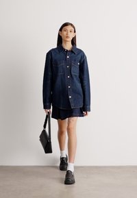 Τζιν πουκάμισο σε σκούρο μπλε, με δύο τσέπες στο στήθος, άνετη εφαρμογή, κλείσιμο με κουμπιά, φορεμένο πάνω από μια ταιριαστή τζιν φούστα, σε συνδυασμό με μαύρα παπούτσια και τσάντα.